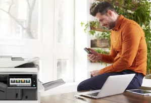 Man using laptop and mobile phone with a printer in foreground